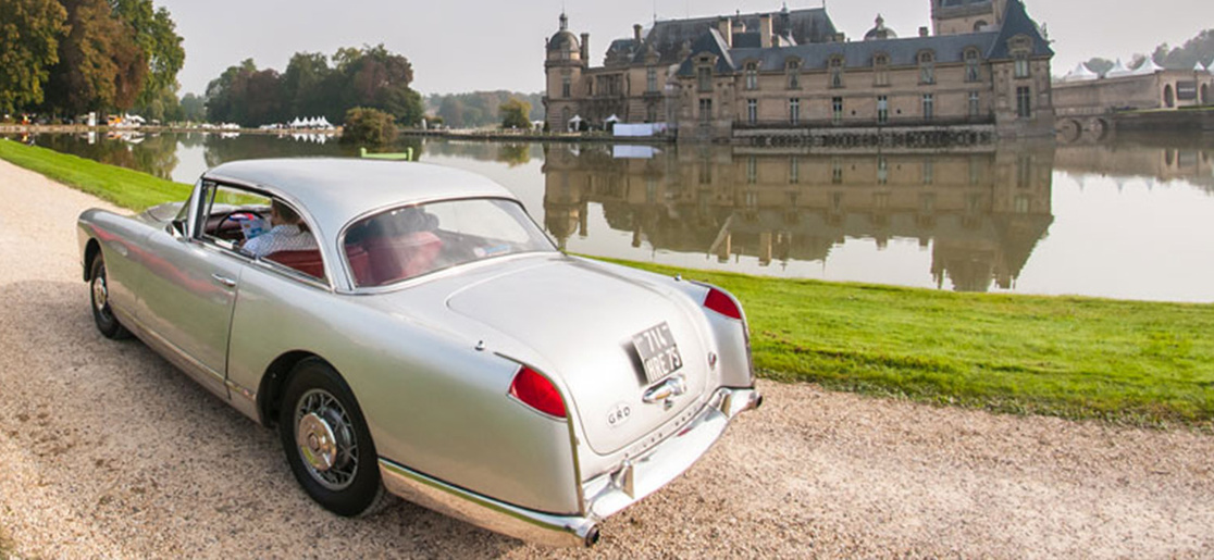 A classic silver car passes through the grounds of a large manor house and its moat