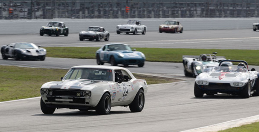 Several cars race around the Daytona Speedway, led by a white car with the number 29 on the side