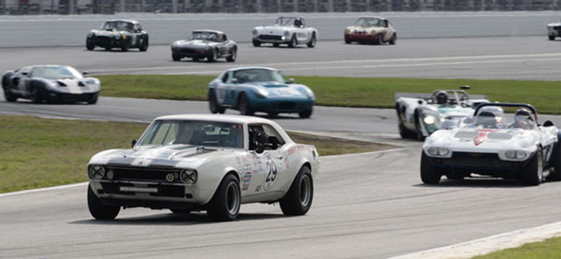 Several cars race around the Daytona Speedway, led by a white car with the number 29 on the side
