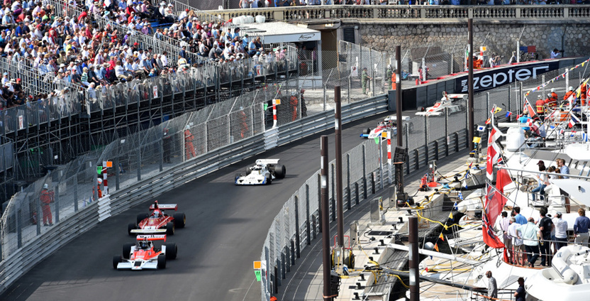 Five cars led by a classic McLaren Formula 1 car race through the Tabac corner in Monaco in front of the yacht harbour and a grandstand filled with fans