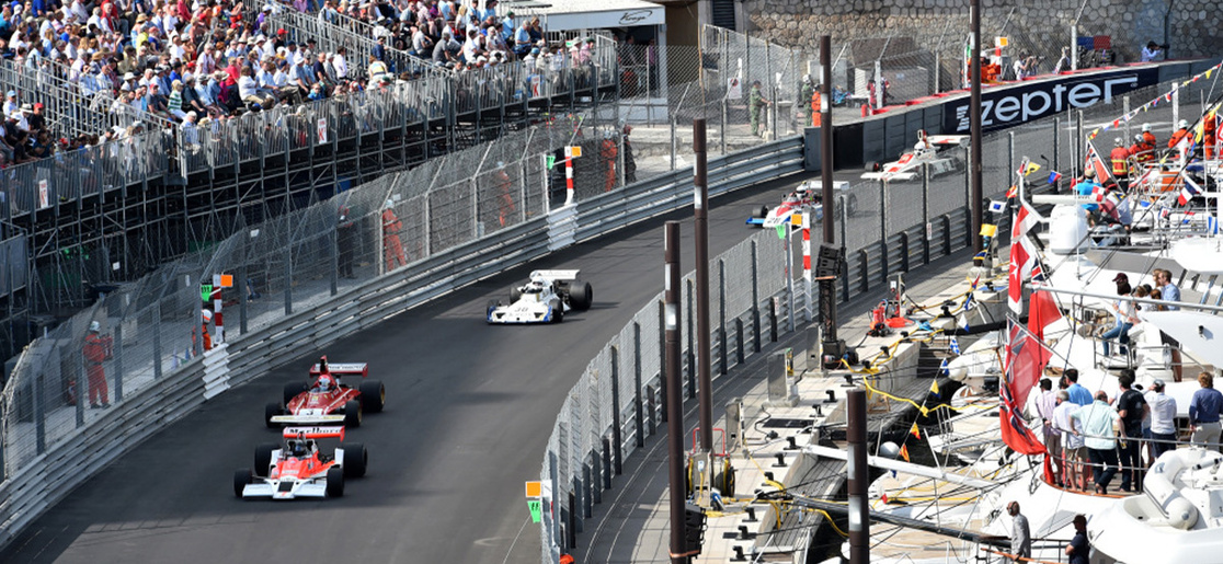 Five cars led by a classic McLaren Formula 1 car race through the Tabac corner in Monaco in front of the yacht harbour and a grandstand filled with fans